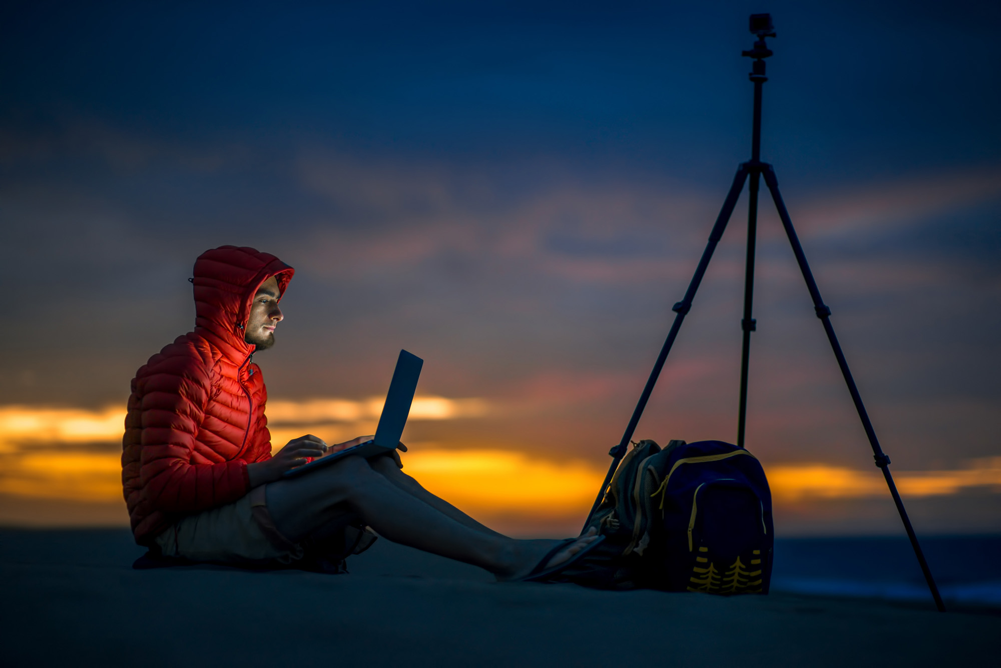 man sat in red jacket editing pictures on laptop night time sunset with tripod